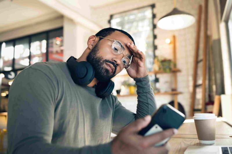 Worried man with glasses and headphones resting his hand on his forehead while looking at his smartphone in a café.
