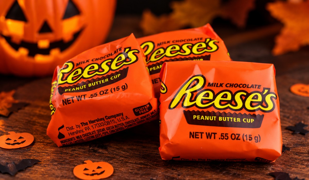 Three Reese’s peanut butter cups in orange wrappers on a dark wooden table with a Halloween pumpkin and autumn decorations.