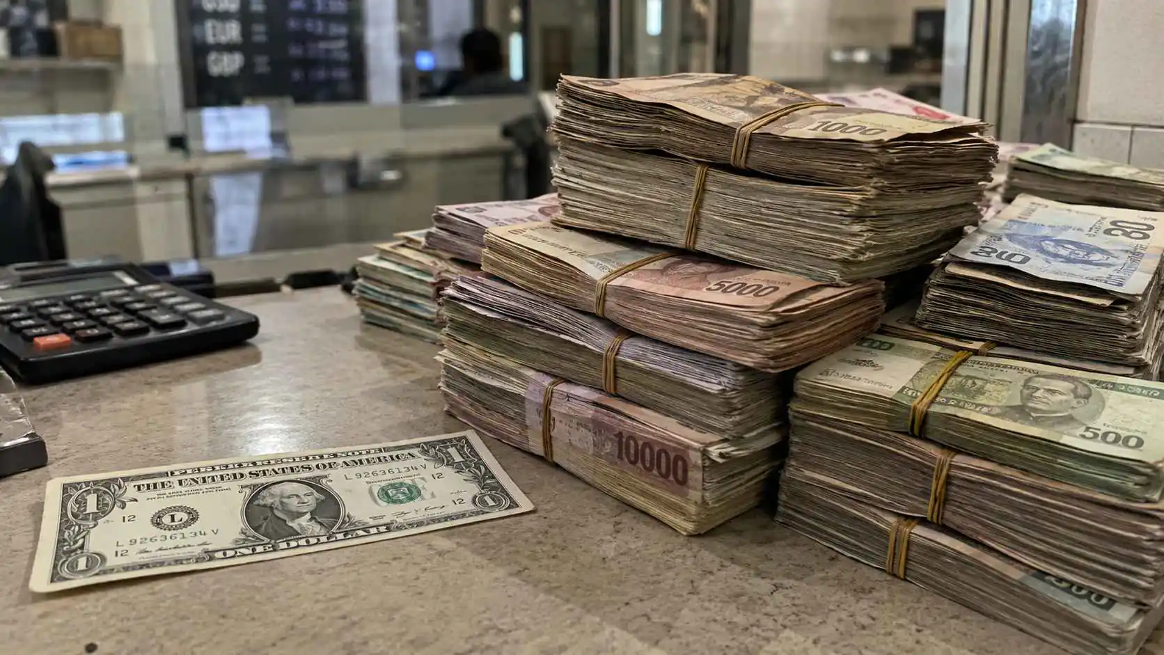 Stacks of low-value foreign currencies beside a single US dollar at a currency exchange counter